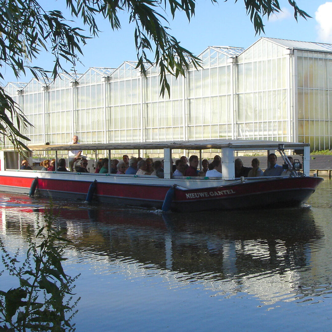 Rode rondvaartboot van Rondvaartbedrijf De Gantel met groep mensen aan boord die langs glazen kassen varen in het Westland