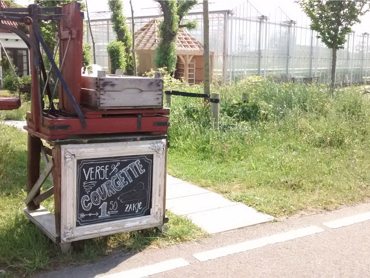 Houten stalletje langs de weg in het Westland met een zwart krijtbord waarop courgettes te koop staan met in de achtergrond glazen kassen in het Westland