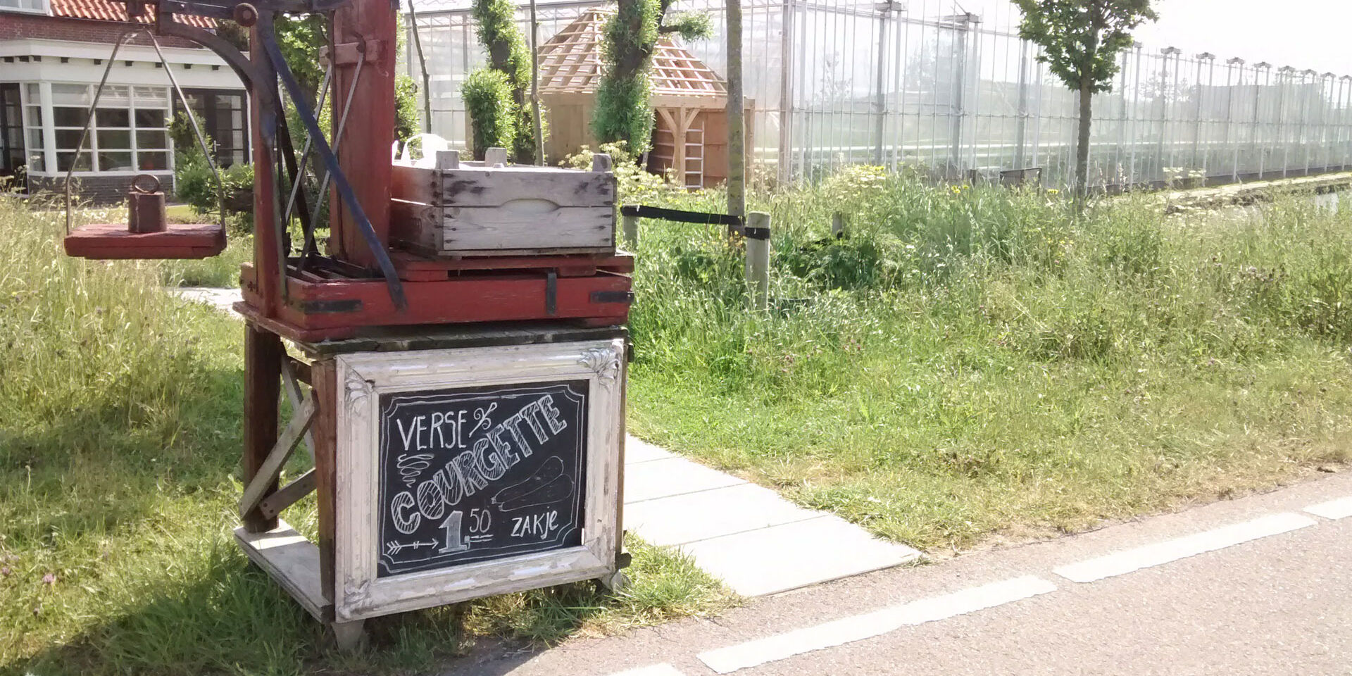 Houten stalletje langs de weg in het Westland met een zwart krijtbord waarop courgettes te koop staan met in de achtergrond glazen kassen in het Westland