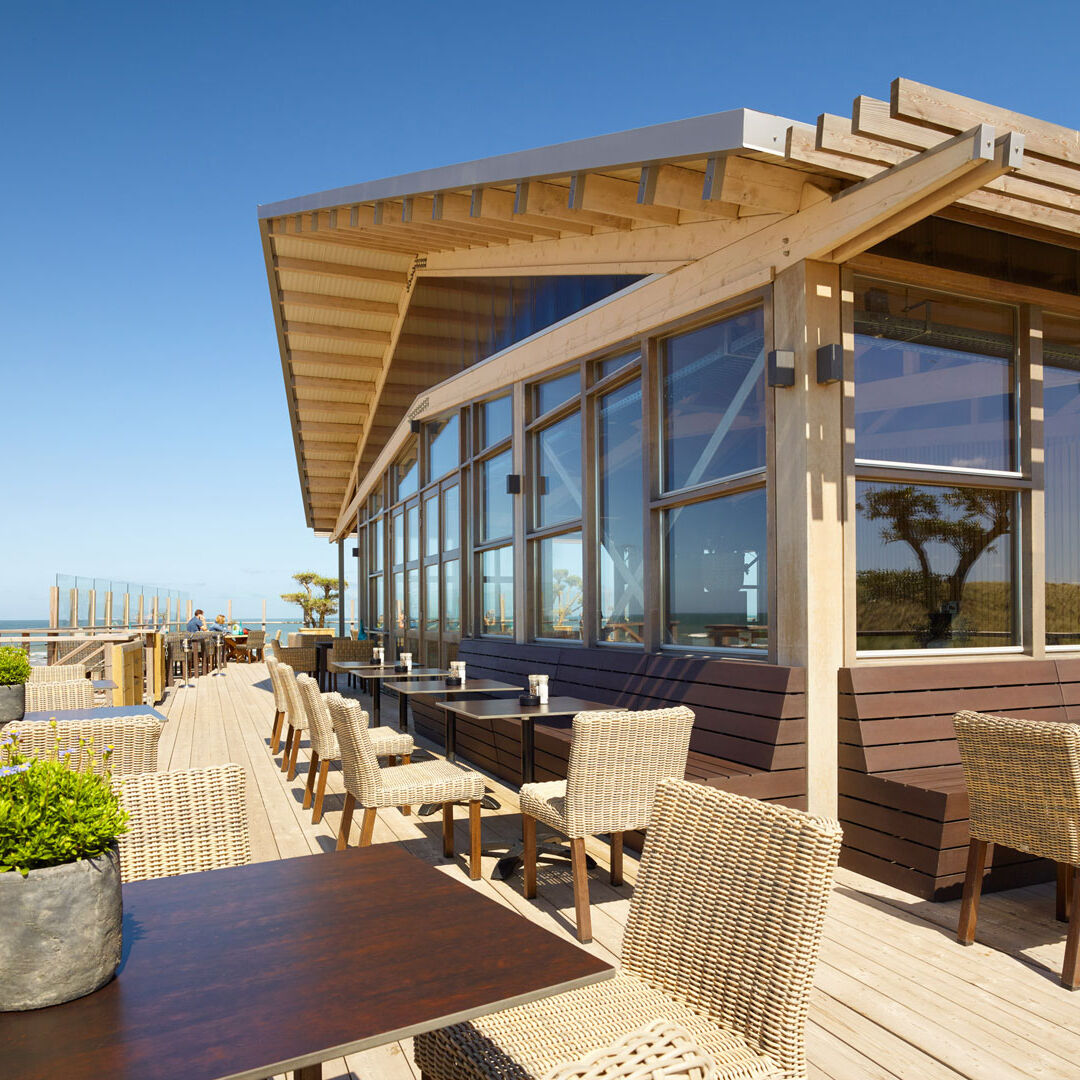 Hooggelegen terras van Restaurant The Coast in Monster met houten vloer, rieten stoelen en glazen gebouw met houten balken dak