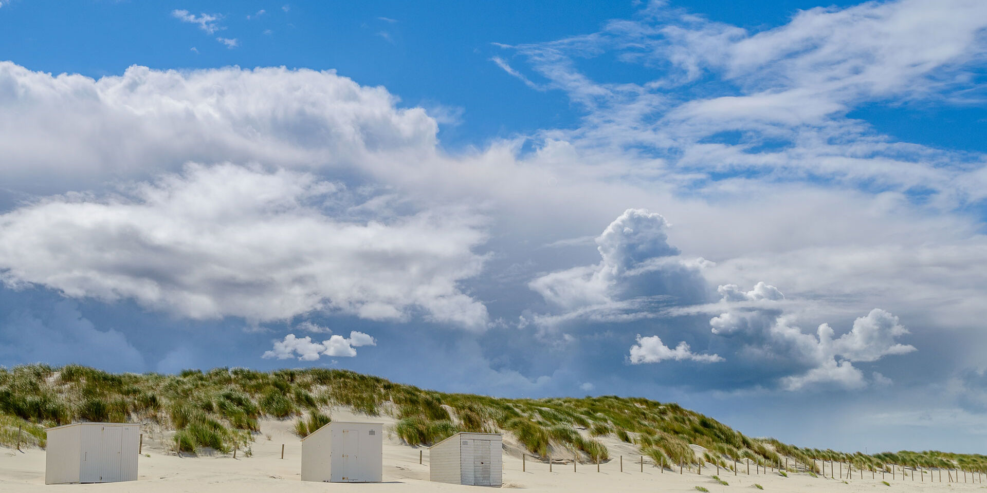 Witte strandhuisjes op het witte zand van het strand in het Westland met op de achtergrond groen begroeide duinen met een blauwe lucht en witte wolken in de zomer