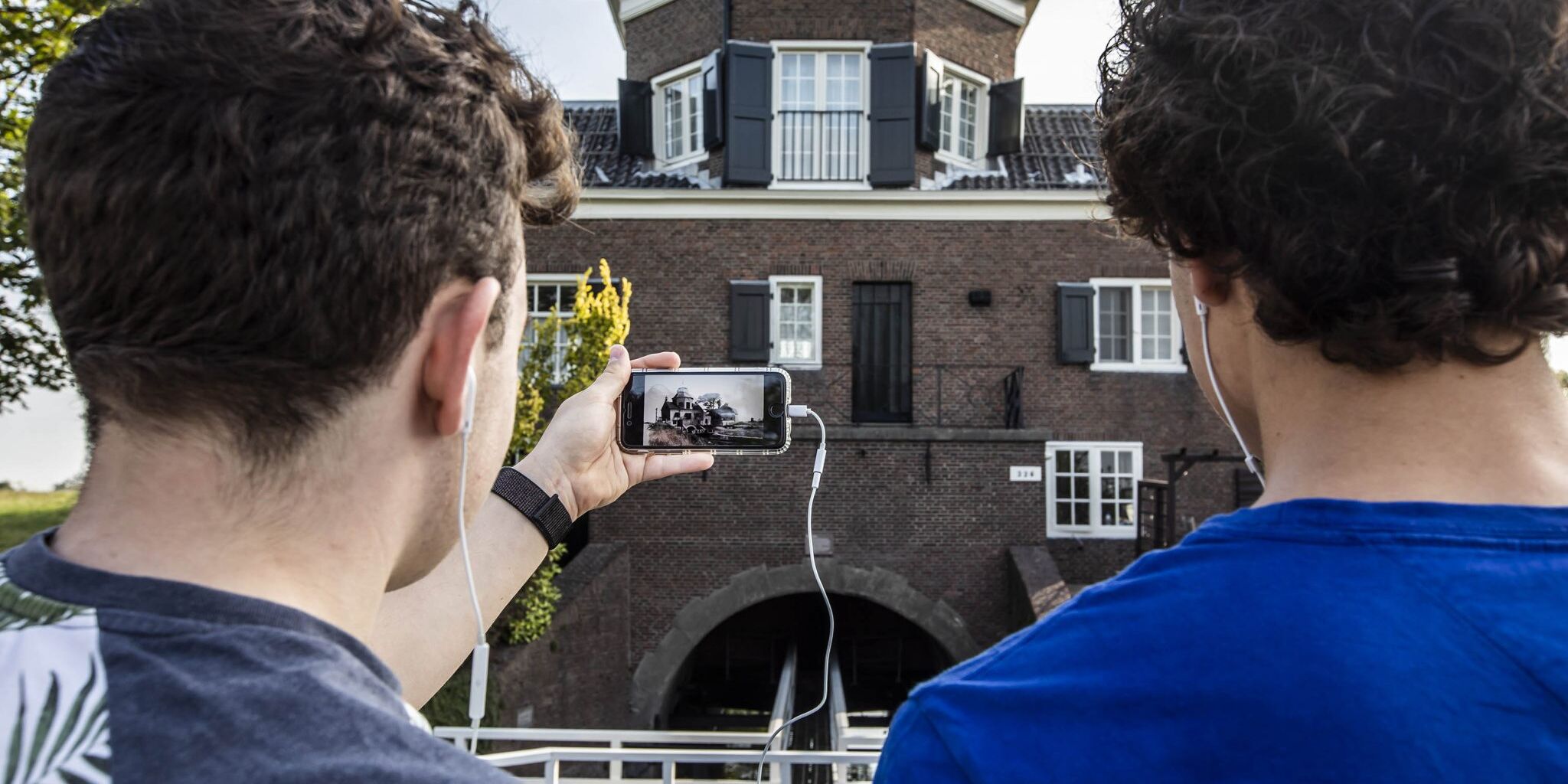 twee mannen staan voor een monument in het Westland en kijken op hun mobiele telefoon naar een video over dat monument tijdens een wandelroutes in het teken van 75 jaar vrijheid in het Westland