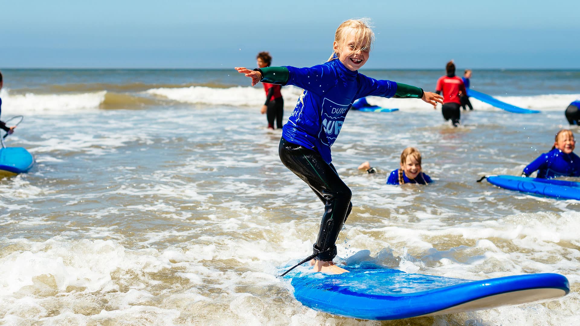 jong meisje met blond haar staat op een blauwe surfplank op zee tijdens een cursus golfsurfen van Dutch Surf Academy in het Westland