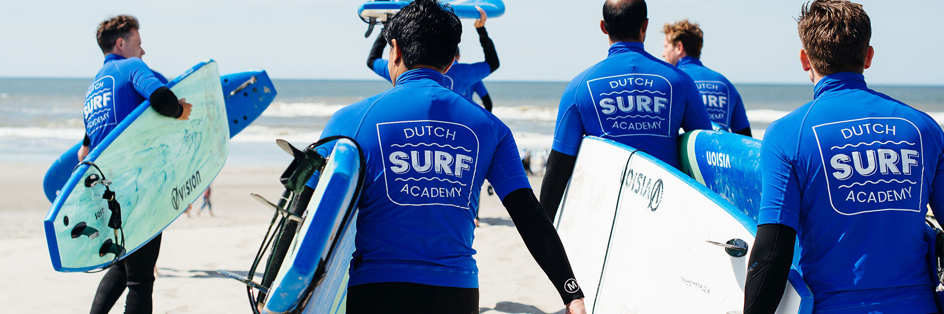 grosp sufers met zwarte wetsuits en blauwe t-shirt lopen op het strand met surfboards onder de arm richting de zee in het Westland