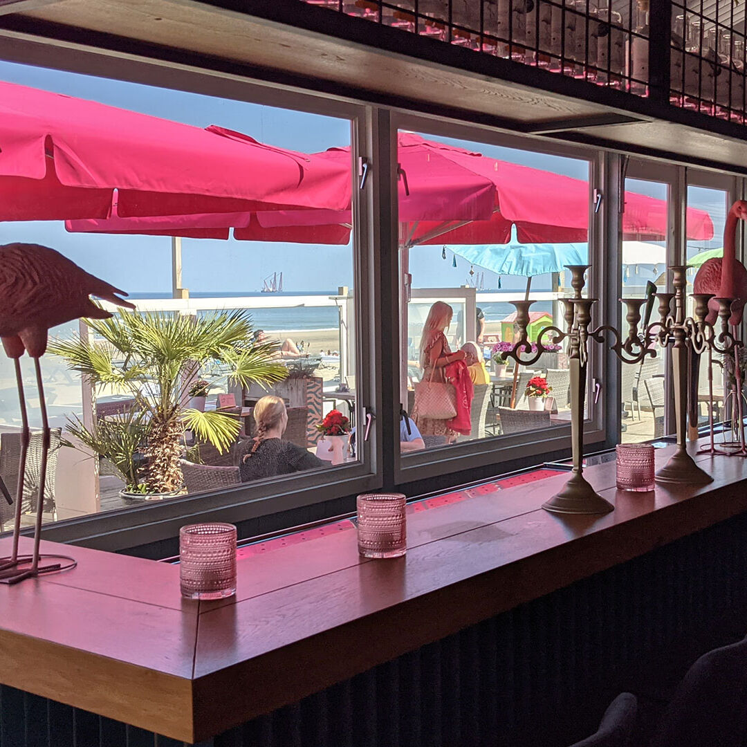 bar in feestzaal met ramen en uitzicht op het buitenterras met roze parasols en de zee
