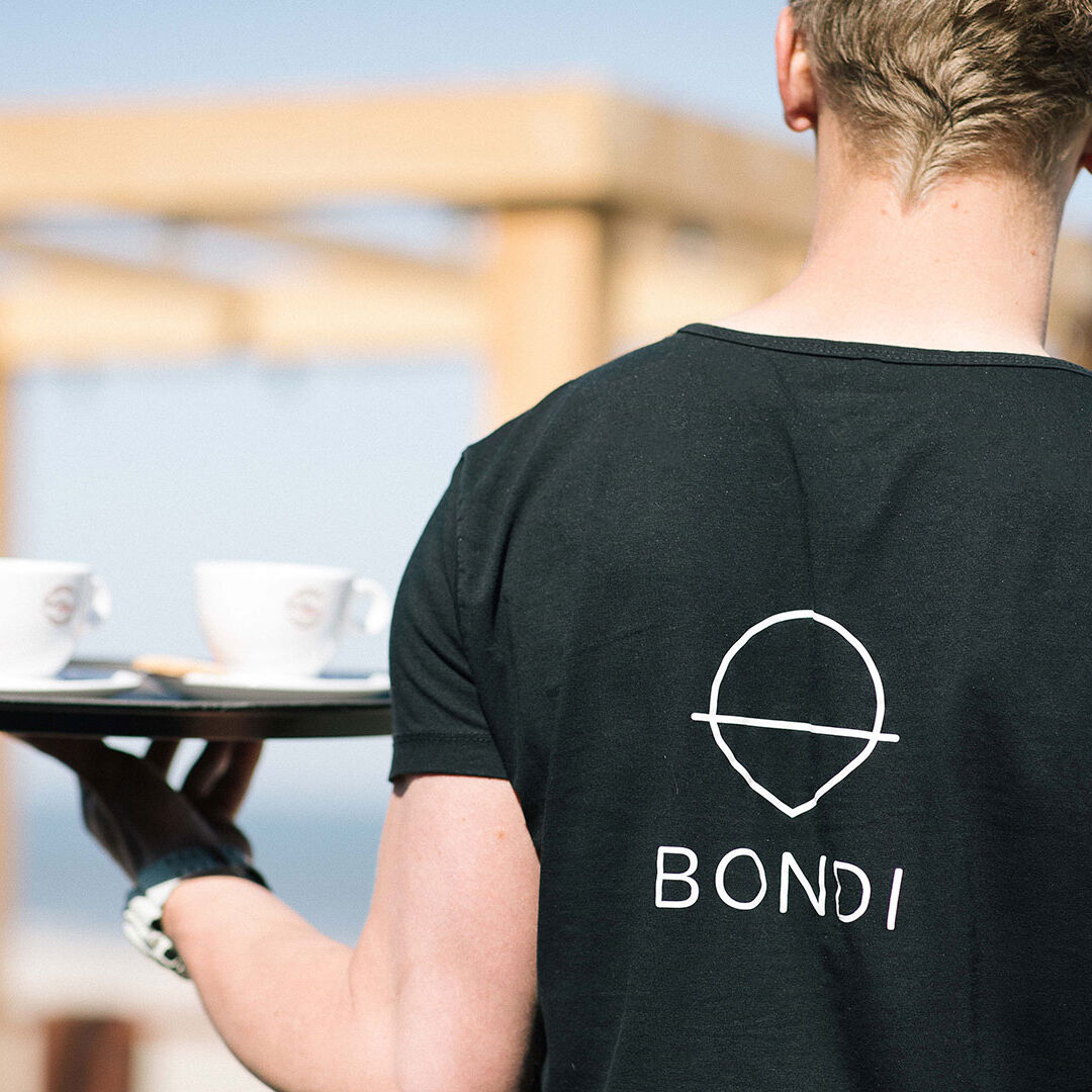 ober met zwart t-shirt met het logo van Bondi Beach Club in Monster in het Westland loopt met een dienblad met 2 koffie over het terras op een zonnige dag aan het strand