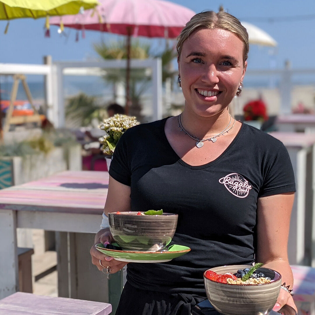 jongedame in zwart t-shirt met logo van Strandtent Bugaloe Beach met 2 bakjes met salade in de hand op het terras van strandtent Bugaloe Beach in het Westland