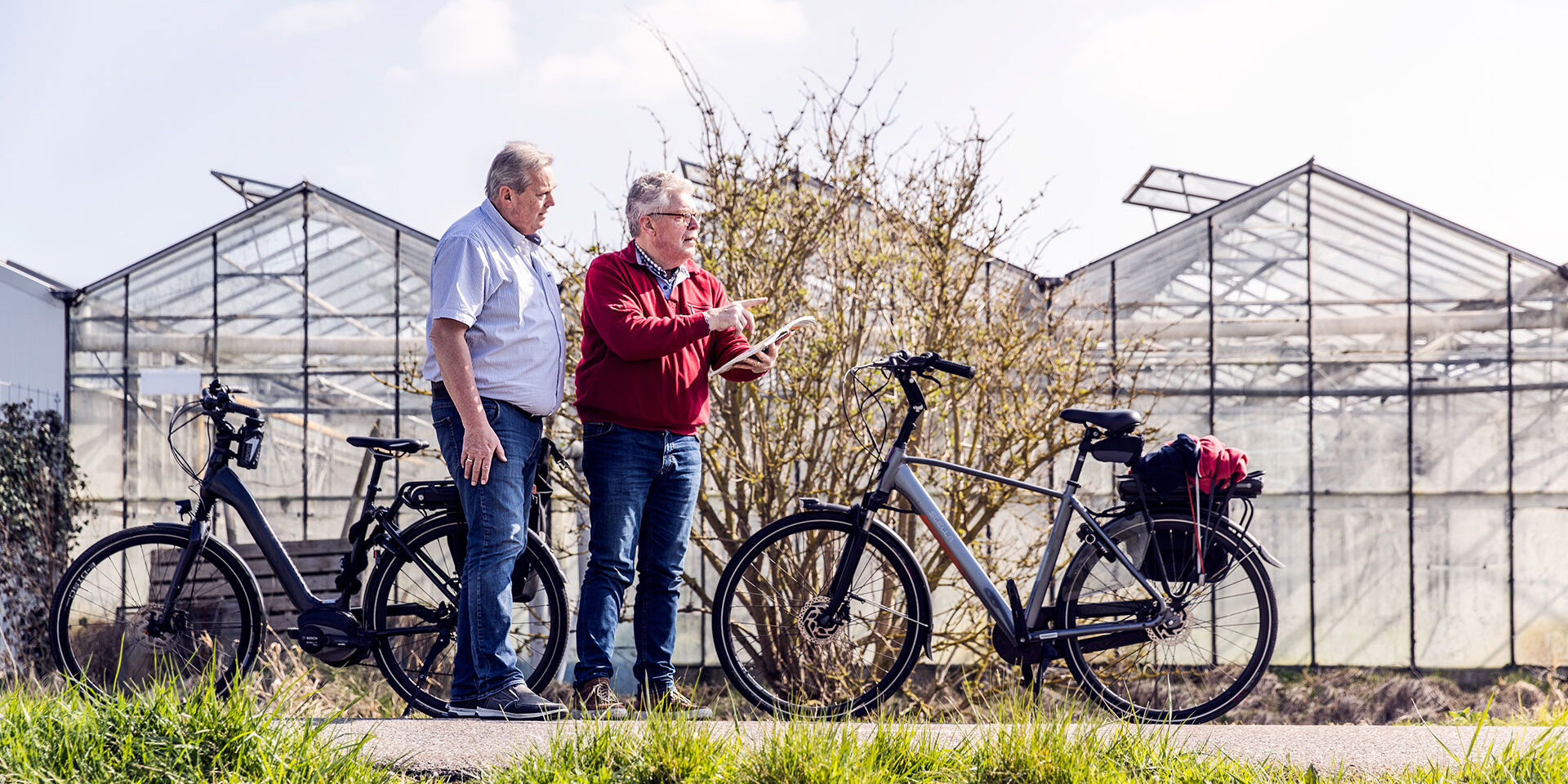 twee fietsende 50Plussers mannen staan naast hun ebike te kijken naar een kaart met de fietsroute met in de achtergrond oude glazen kassen langs een fietspad in het westland op een zonnige dag