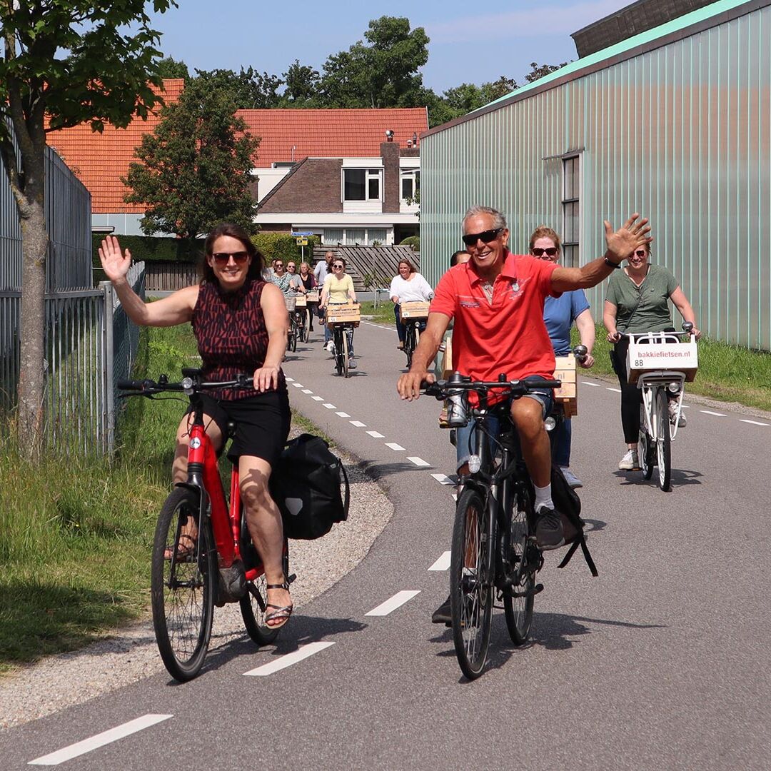 groep fietsers op fietsen met een houten kratje voorop (Bakkie Westland) tussen de kassen van het Westland tijdens een Bakkie Fietsen fietstour