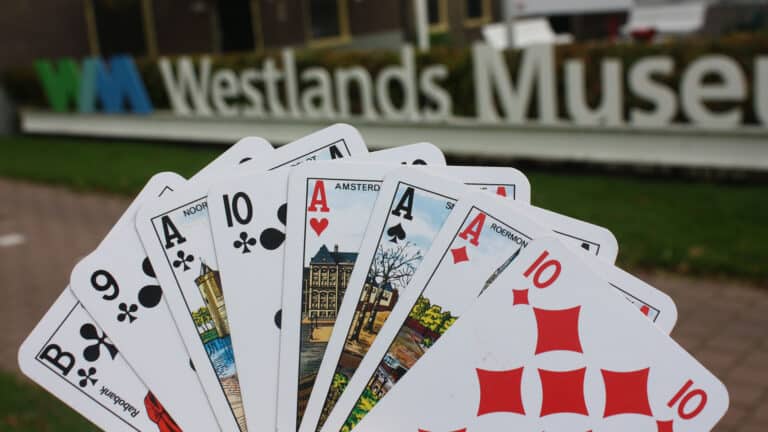 hand met speelkaarten in de hand met op de achtergrond het Westlands Museum als aankondiging van klaverjas wedstrijden in het museum in de maanden oktover t/m april