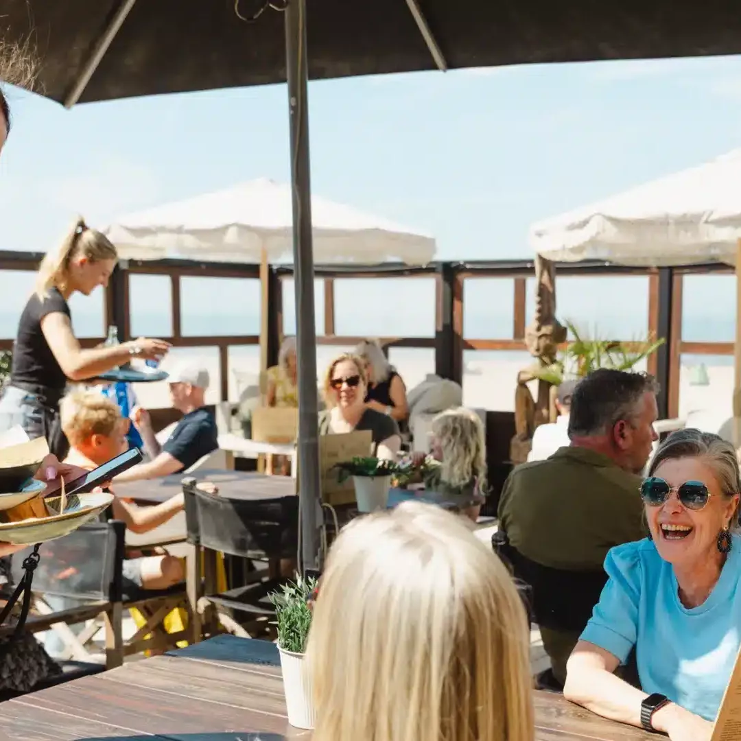 twee gasten op het terras van een strandtent in het Westland bestellen iets te eten bij een jonge dame op een zonnige dag aan de kust