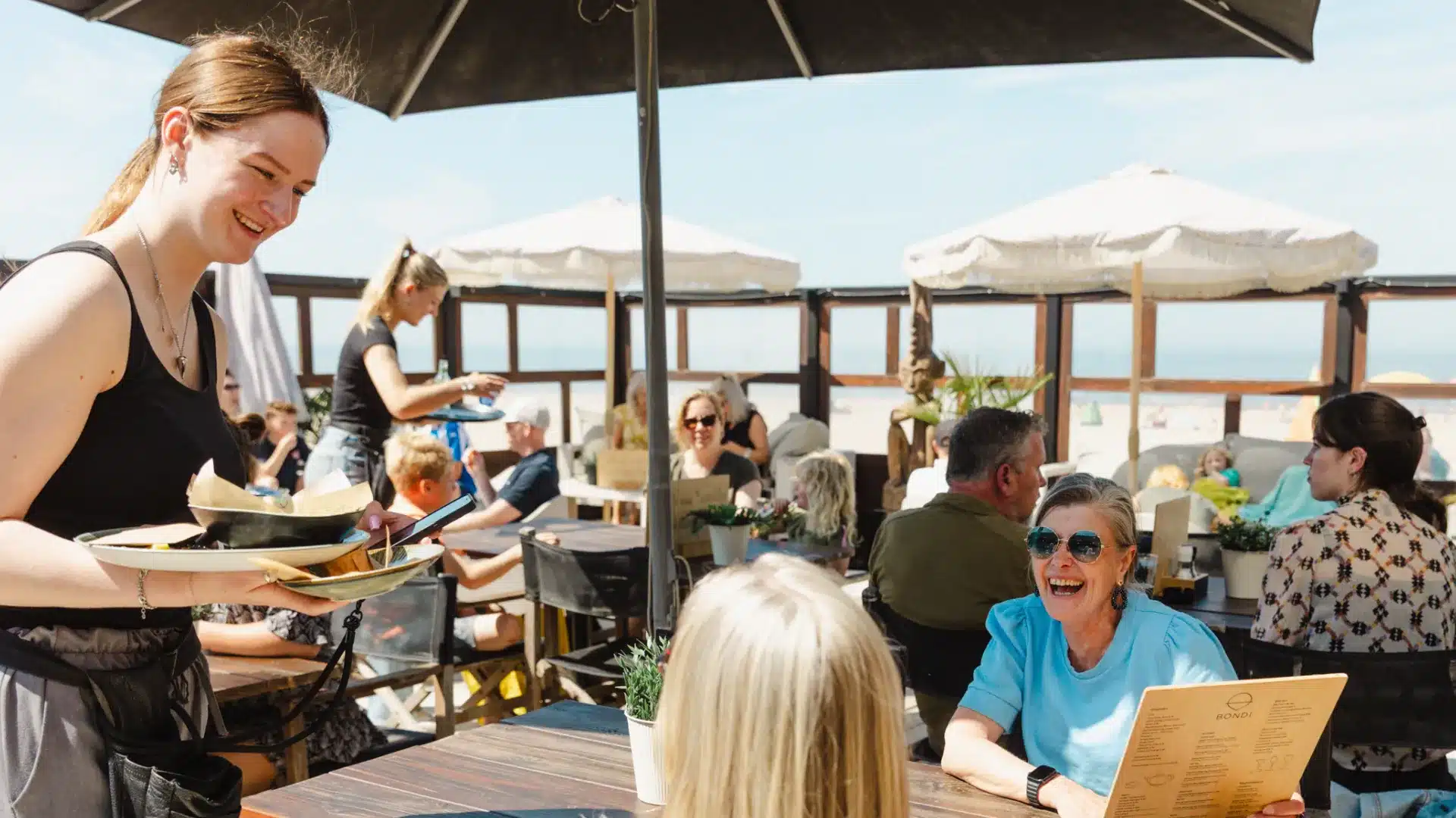 twee gasten op het terras van een strandtent in het Westland bestellen iets te eten bij een jonge dame op een zonnige dag aan de kust
